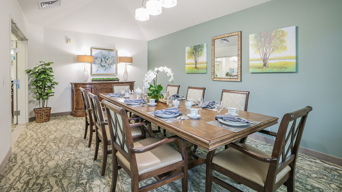 A dining room with a wooden table set for eight, surrounded by chairs. The table has a white orchid centerpiece. The walls are green with two tree paintings and a sideboard with lamps and a potted plant.