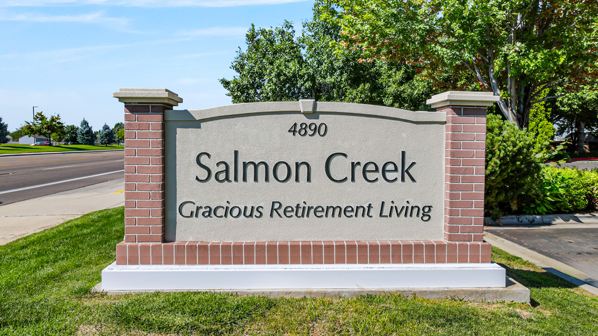 A stone and brick sign reads "4890 Salmon Creek Gracious Retirement Living," surrounded by grass, trees, and a road on a sunny day.