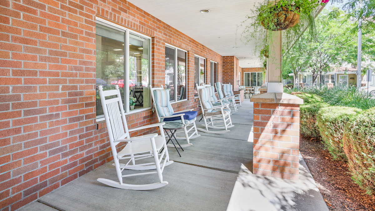 A row of white rocking chairs lines a covered outdoor patio with brick walls and large windows, overlooking greenery and hanging plants.