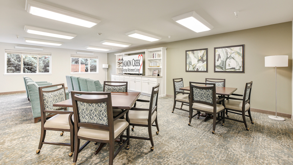 A bright, spacious room with two tables and chairs, teal upholstered armchairs, large windows, framed artwork, and a white bookshelf with a "Salmon Creek" sign on the back wall. Ceiling lights illuminate the area.