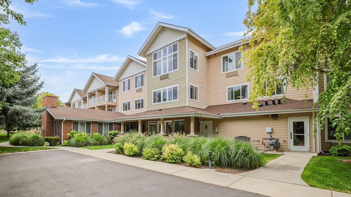 A three-story beige apartment building with large windows, surrounded by green trees and shrubs, under a blue sky. A sidewalk and driveway run along the front entrance.