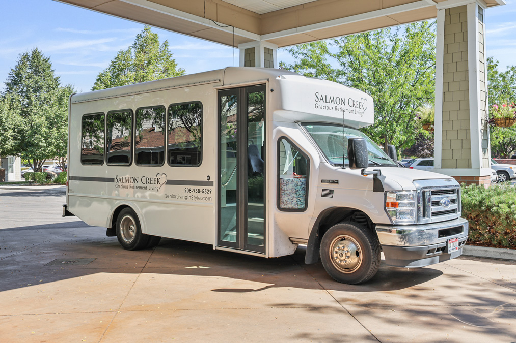 A white Salmon Creek Retirement Living shuttle bus is parked under a covered entrance, surrounded by trees and greenery on a sunny day.