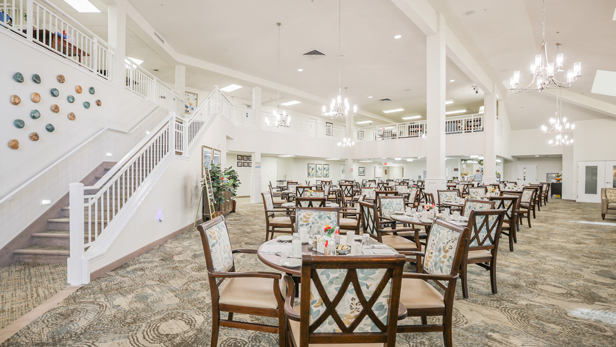 A spacious, well-lit dining area with multiple round tables and cushioned chairs, a carpeted floor, chandeliers, wall art, and a staircase leading to an upper level in a modern building.