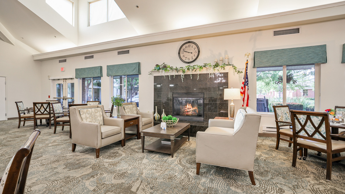 A bright, spacious lounge with armchairs, a coffee table, and a fireplace beneath a large clock. Windows line the walls, and an American flag stands beside the fireplace. The room has neutral colors and decorative greenery.