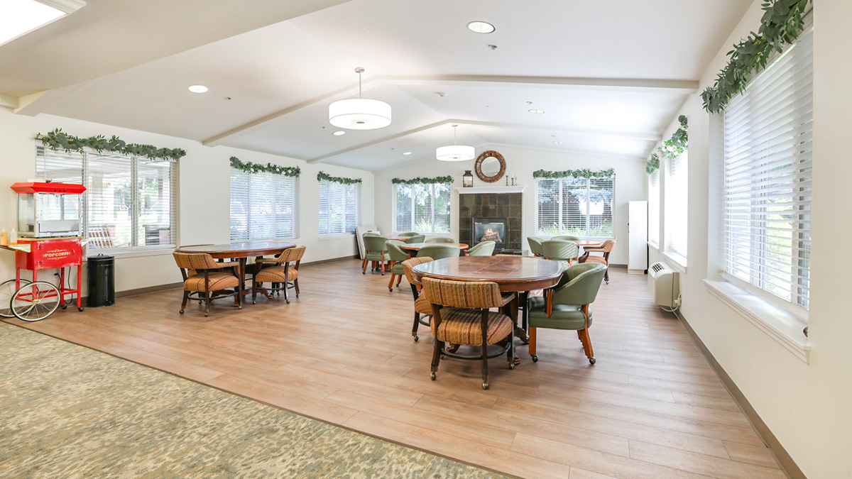 A bright, spacious community room with wooden floors, several round tables with chairs, green armchairs near a fireplace, large windows with white blinds, and a red popcorn machine in the corner.