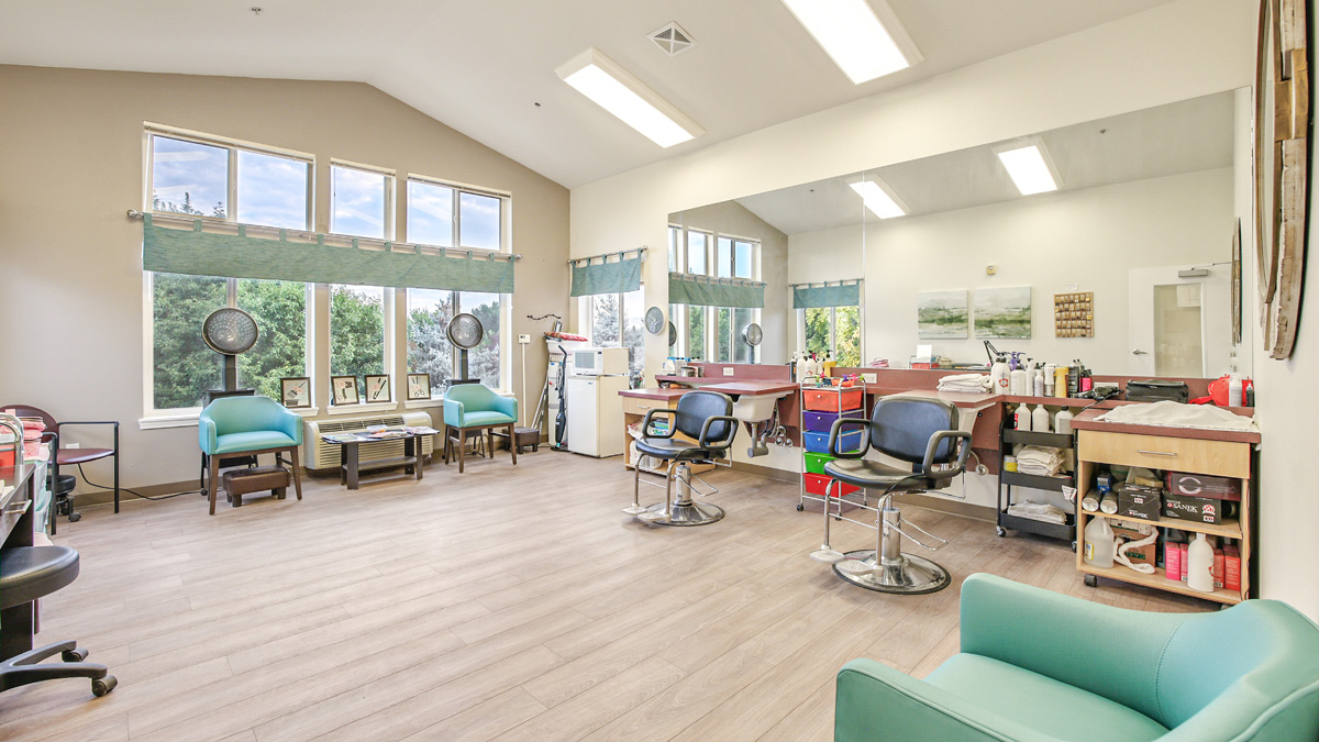 Bright, spacious salon with large windows, wooden floors, styling chairs, mirrors, hair dryers, and shelves stocked with hair products and tools. Natural light fills the modern, tidy room.