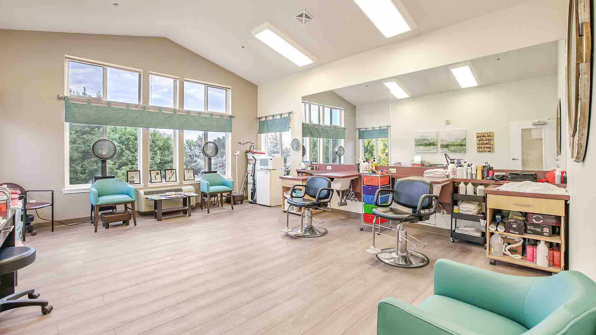 A bright, modern salon with large windows, mirrors, and salon chairs. Hairdryers are positioned by the windows, and shelves hold hair products and supplies. The space is clean and well-lit with natural and overhead lighting.