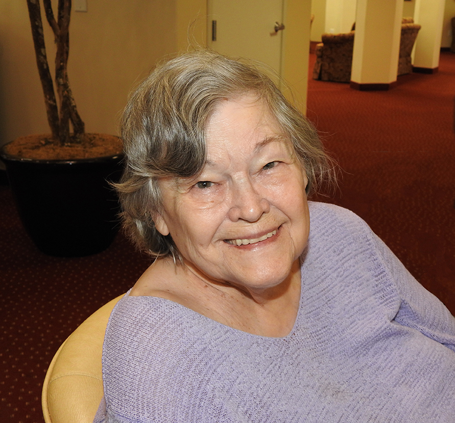 An elderly woman with gray hair is smiling warmly. She is wearing a light purple knitted sweater and is seated indoors. The background shows a red-carpeted floor, a large plant in a black pot, and some furniture.