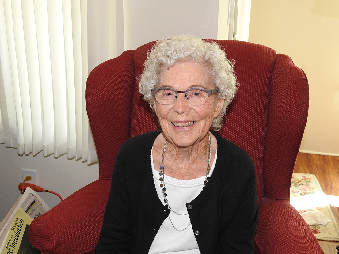 An elderly woman with curly white hair and glasses smiles while sitting in a red armchair. She is wearing a black sweater over a white shirt and a beaded necklace. Sunlight filters through vertical blinds behind her, and a magazine is partially visible next to the chair.
