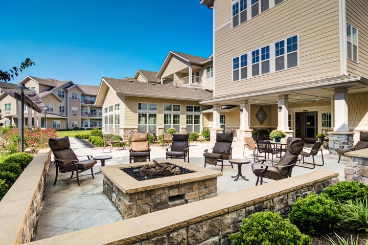 An outdoor patio at a modern residential complex features cozy seating around a central fire pit. The beige buildings have large windows and neatly maintained gardens, creating a welcoming and serene environment under a clear blue sky.