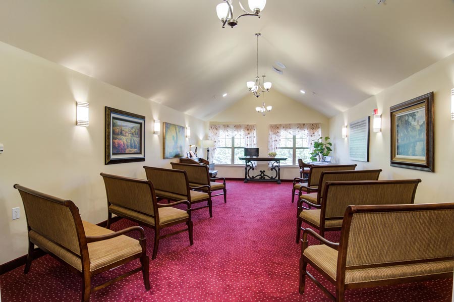 A room with a red carpet, wooden benches with cushions arranged in rows, paintings on the walls, a piano at the far end, and large windows with light curtains. Hanging light fixtures are on the ceiling, and a potted plant is near the windows.