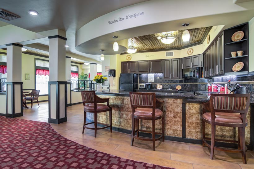 A spacious, well-lit hotel dining area with a bar. The bar has three cushioned high chairs and a granite countertop. The kitchen behind the bar has dark wooden cabinets. The dining area features tables and chairs, with large windows and red curtains.