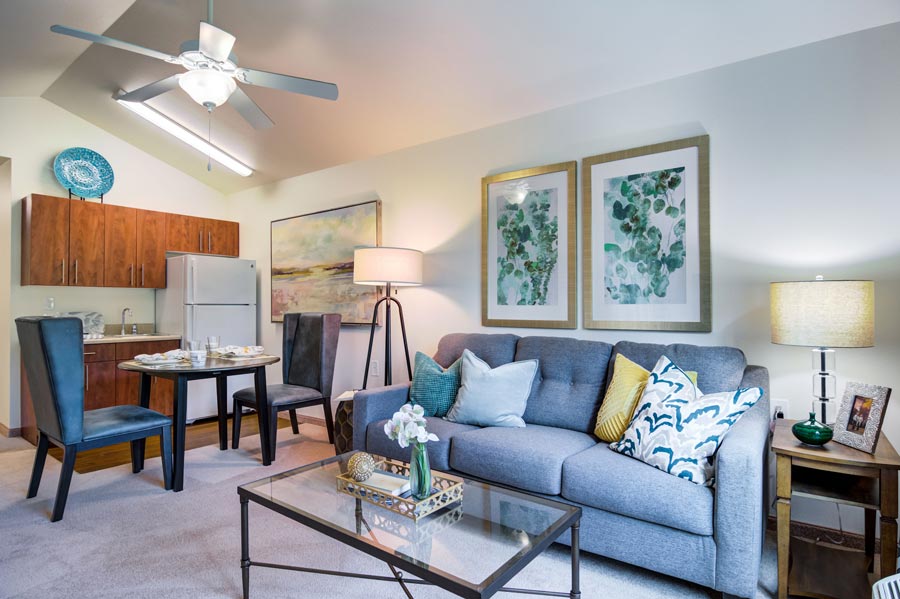 A cozy living room features a gray sofa adorned with colorful throw pillows, a glass coffee table, and a floor lamp. Behind, there's a small dining area with a round table and two chairs. The kitchenette has wooden cabinets. Art pieces and a ceiling fan complete the decor.