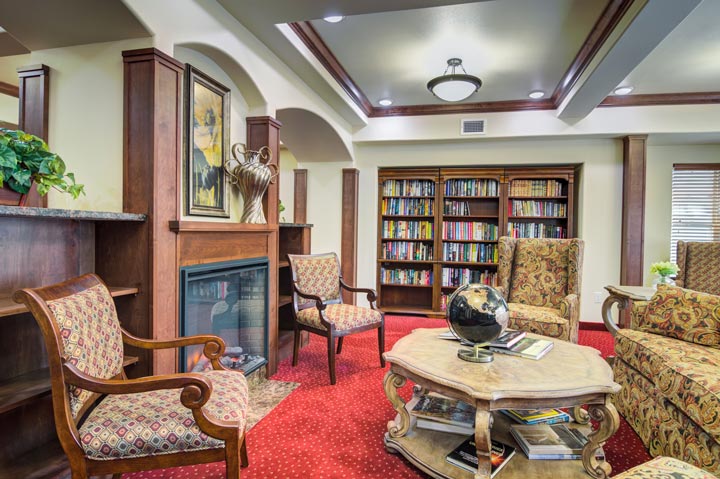 A cozy library room with red carpeted flooring, wooden bookshelves filled with books, and upholstered armchairs. A wooden coffee table with a decorative globe, books, and a floral arrangement sits in the center. A fireplace is surrounded by elegant decor.