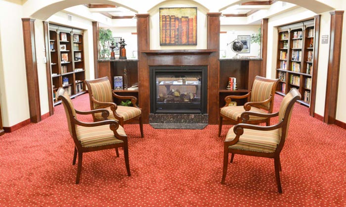 A cozy library setting features four wooden armchairs with patterned cushions arranged in a semicircle on a red carpet with white dots in front of a fireplace. Book-filled shelves line the walls, and plants add a touch of greenery. A painting hangs above the mantle.