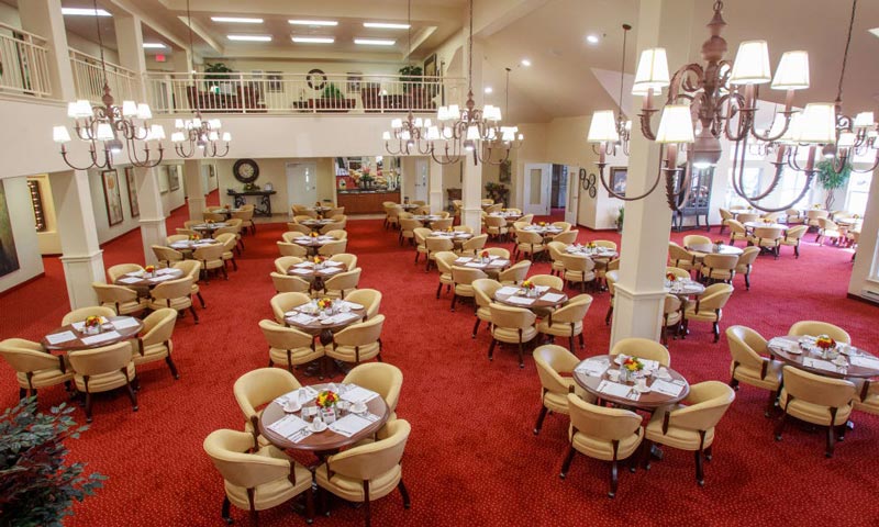 A spacious dining hall with red carpet features several round tables, each set with four beige chairs. Tables are adorned with floral centerpieces and set with napkins. Large chandeliers hang from the ceiling, adding to the elegant ambiance.