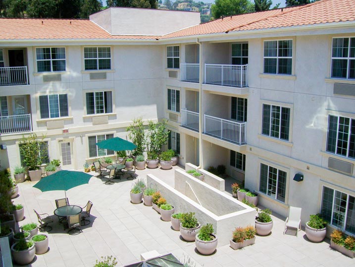 An open courtyard of a residential building with multiple floors and white walls. The area has potted plants, green umbrellas, and outdoor seating with tables and chairs. The courtyard has ample natural light and overlooks balconies and large windows of the apartments.