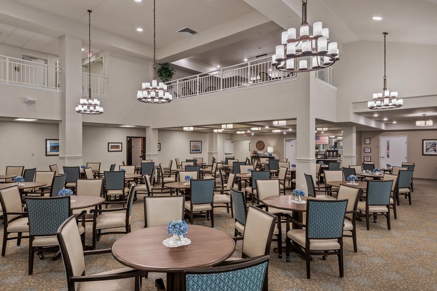 A spacious dining area with neatly arranged tables and chairs. Two chandeliers hang from the ceiling, providing ample light. The tables have small floral arrangements as centerpieces. A balcony overlooks the dining room, and the overall décor is modern and elegant.