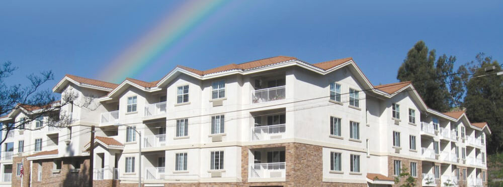 A multi-story residential building with white walls and brownish-tiled roofs is pictured under a clear blue sky. A vivid rainbow arcs across the sky above the building. Trees are visible in the background.