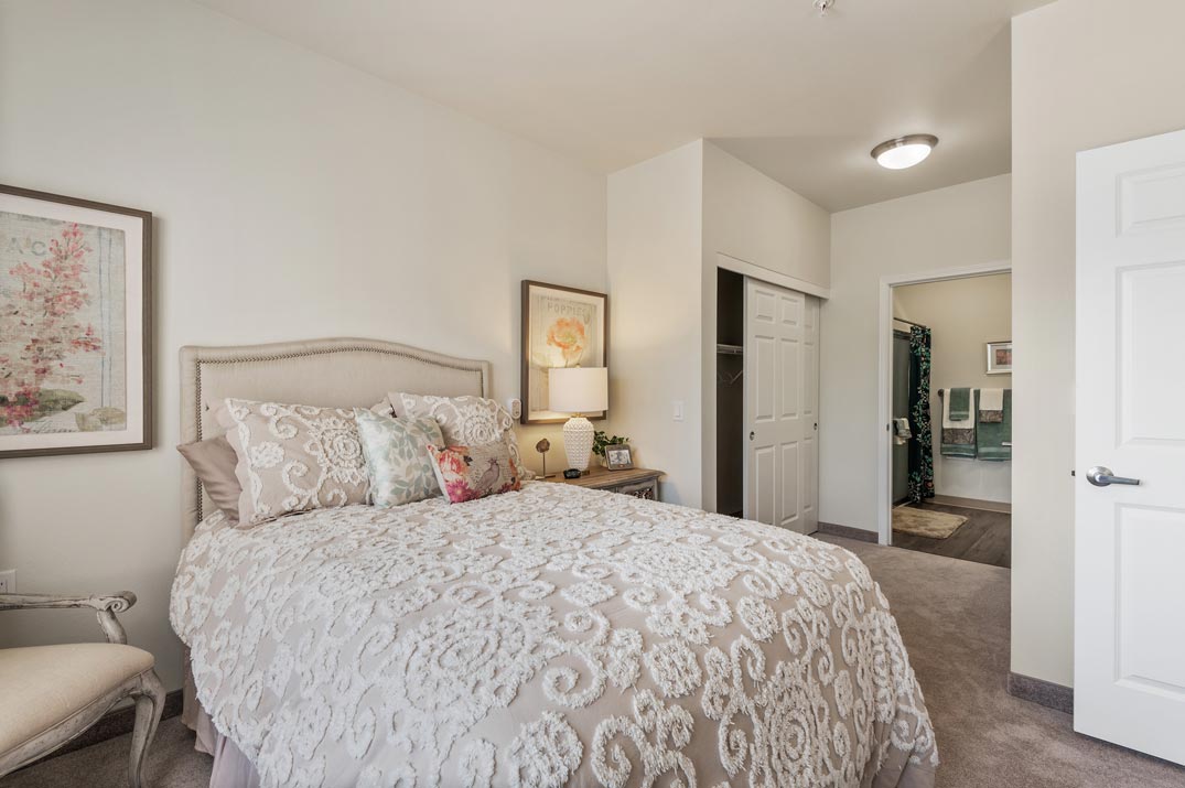 A cozy bedroom featuring a plush bed with a patterned white and beige comforter, pillows, and framed art on the walls. There's a nightstand with a lamp and decor, an armchair to the side, and an open door revealing a closet and a bathroom.