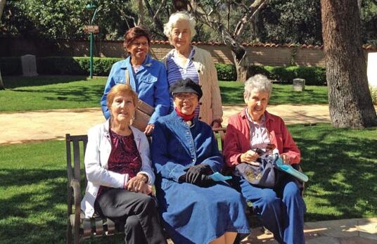 Five elderly women are enjoying a sunny day in a park. Three are sitting on a bench while two stand behind them. The women are smiling, and they appear relaxed and content. The park is lush with greenery and trees in the background.