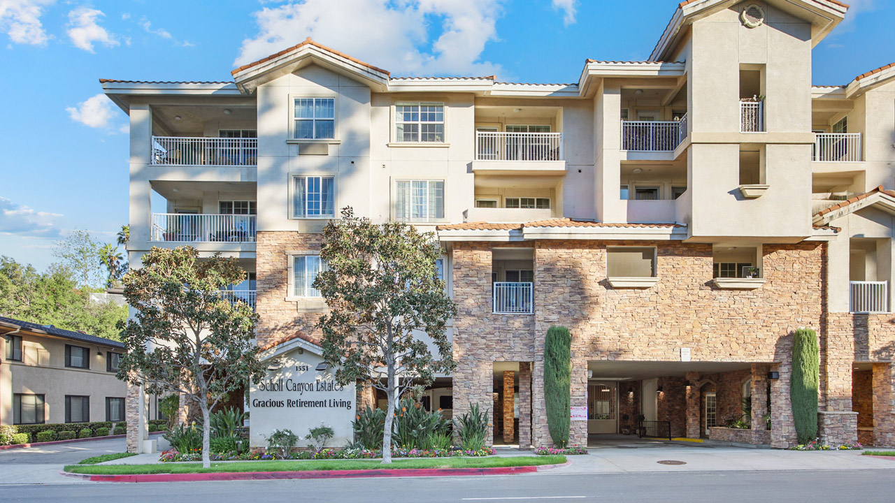 A modern, multi-story retirement living building with balconies, stone façade, and landscaped greenery at the entrance. A sign reads "Gardens Retirement Living" near the driveway under a covered archway.