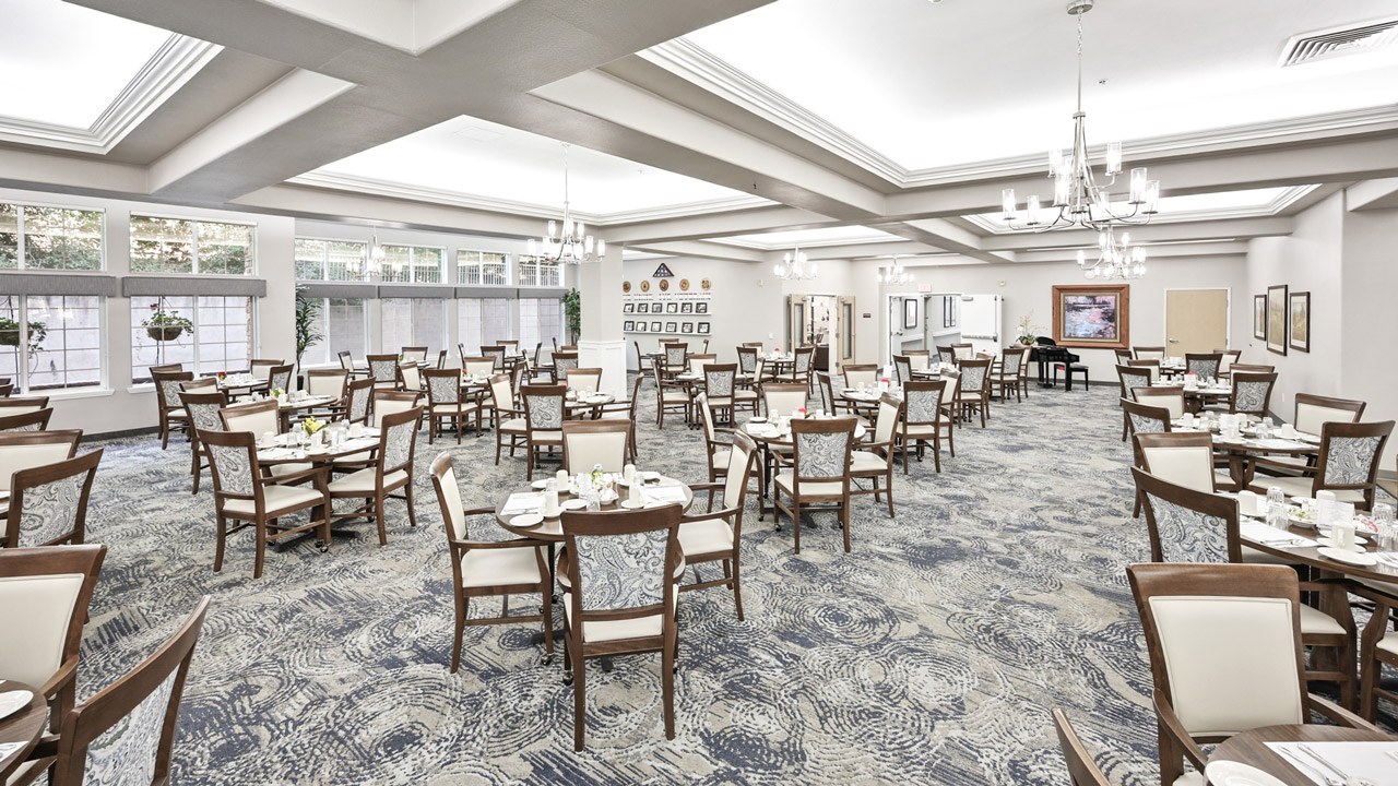 A spacious, well-lit dining room with many neatly arranged tables and chairs, set for a meal. Large windows let in natural light, and elegant chandeliers hang from the ceiling above patterned carpet floors.