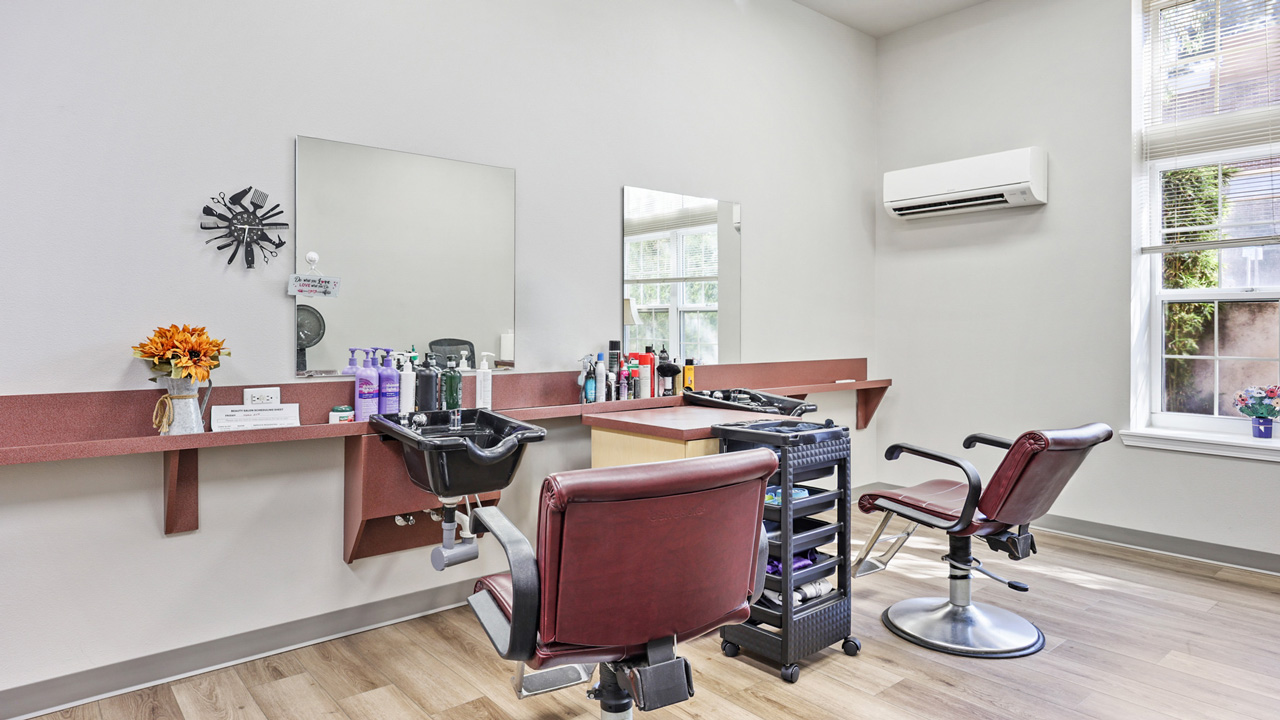 A bright hair salon features two maroon chairs, a washing sink, mirrors, hair products, and a rolling cart, with sunlight streaming in through large windows and a wall-mounted air conditioner.