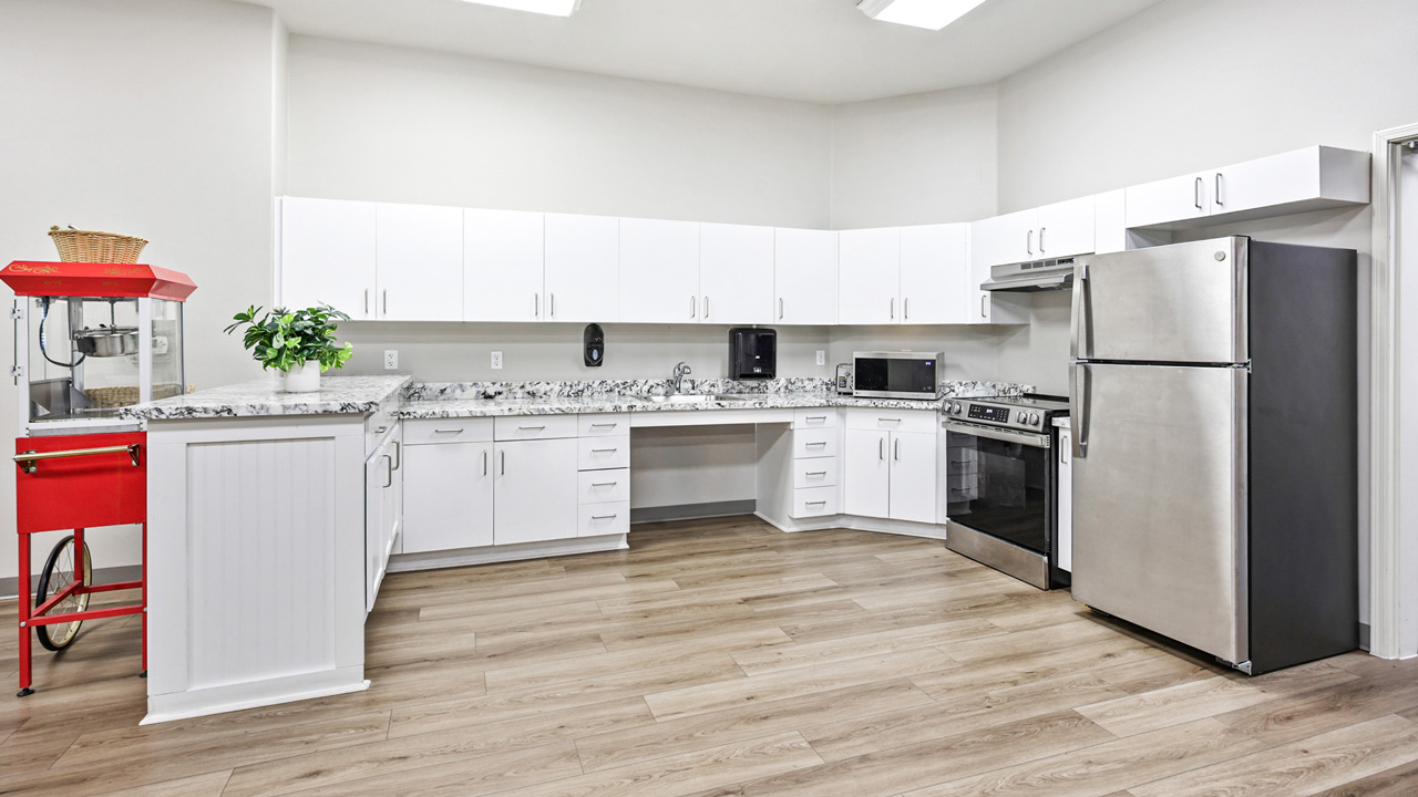 Modern kitchen with white cabinets, stainless steel appliances, marble countertops, light wood flooring, a microwave, a potted plant, and a red popcorn machine on the left side.
