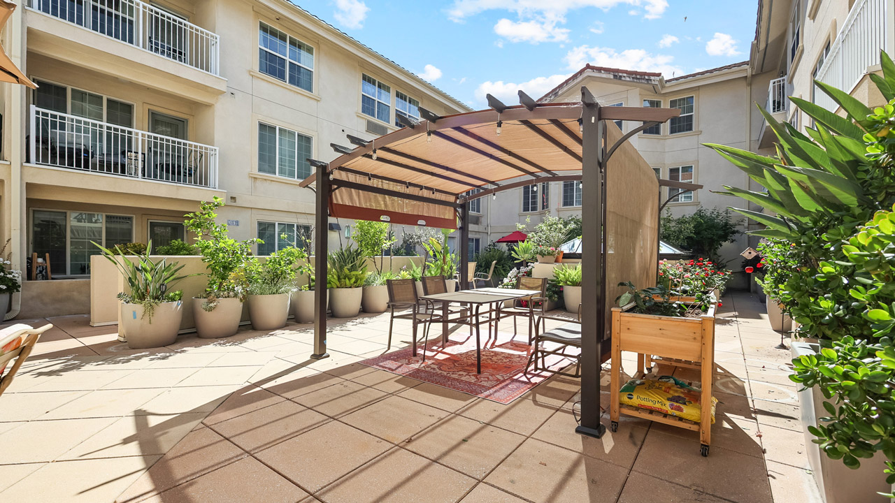 A sunny apartment courtyard with a pergola covering a table and chairs, surrounded by potted plants and greenery. Three-story beige buildings with balconies enclose the space.