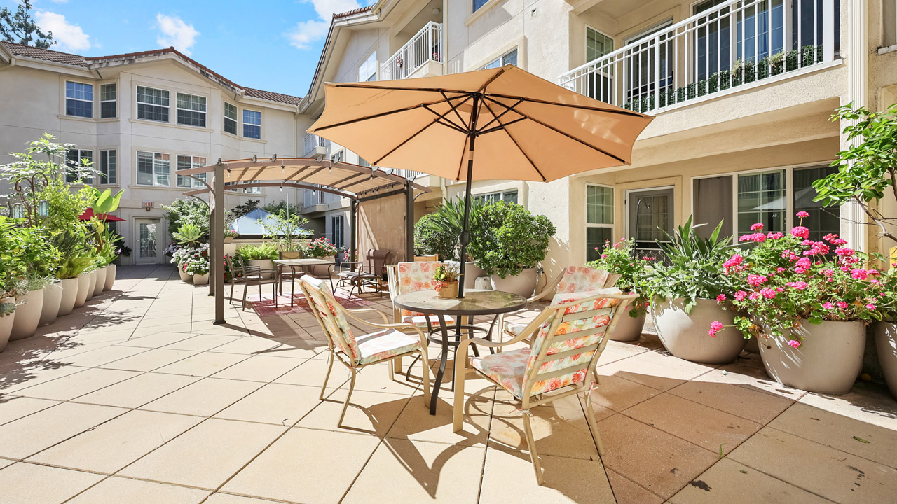 A sunny outdoor patio with a round table, floral cushioned chairs, and a large beige umbrella. Potted plants and flowers line the tiled patio, with apartment buildings and balconies in the background.