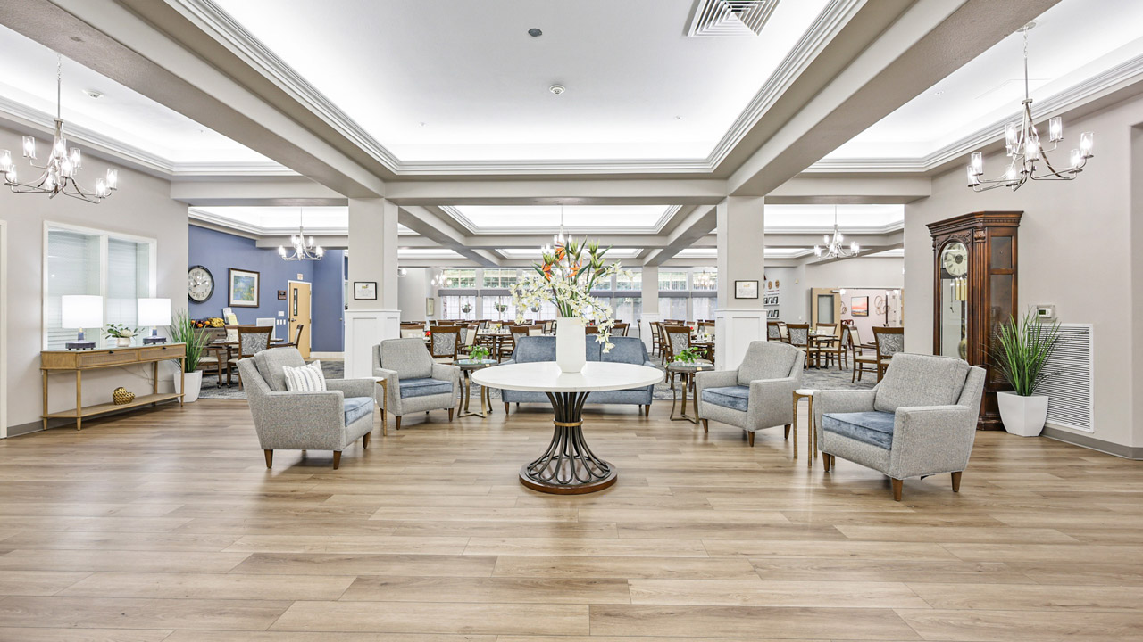 A spacious, modern lobby with light wood flooring, a round table with a flower arrangement in the center, gray armchairs arranged in a circle, and bright overhead lighting. Chandeliers and large windows enhance the airy feel.
