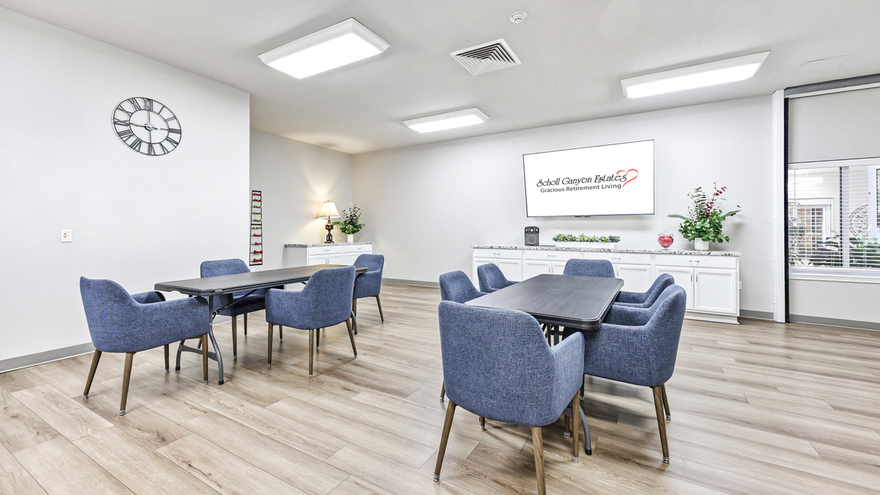 Bright, modern meeting room with three tables, blue upholstered chairs, wood flooring, wall clock, plants, and a large sign reading "Edelweiss Campus Rehabilitation" on the far wall.