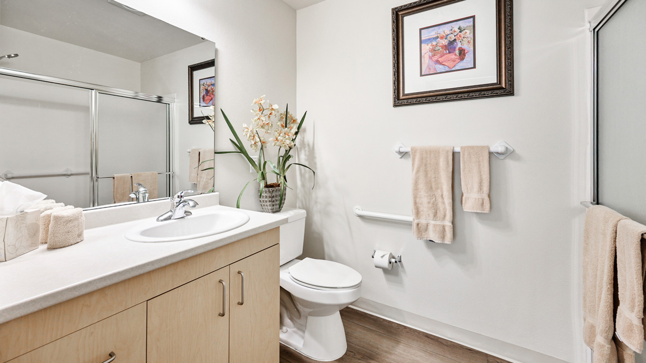 Modern bathroom with light wood vanity, white countertop and sink, toilet, beige towels, a large mirror, potted orchid, framed floral artwork on the wall, and a glass shower door.