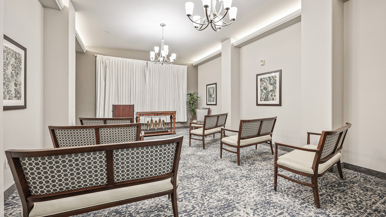 A small, bright room with patterned armchairs arranged in rows facing a wooden table and white curtains, with chandeliers above and framed art on light-colored walls.