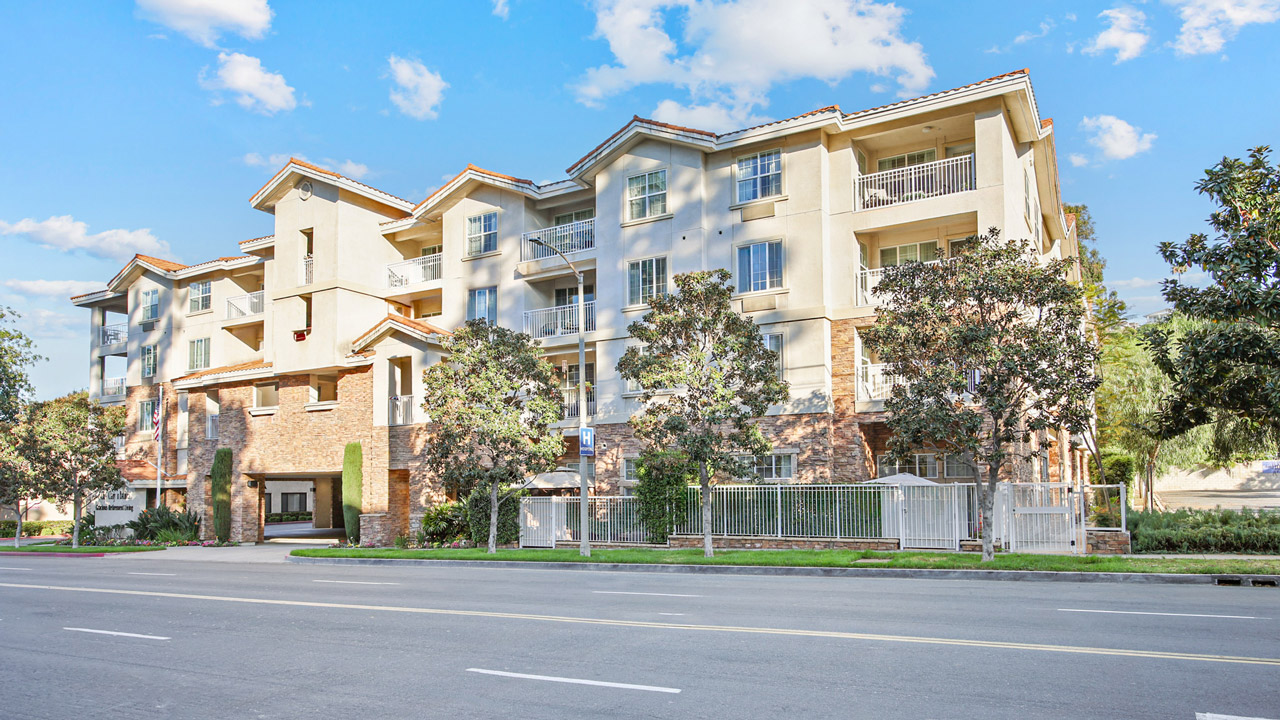 A modern four-story apartment building with beige walls and balconies, surrounded by green trees and a metal fence, situated along a sunny, empty street under a blue sky with scattered clouds.