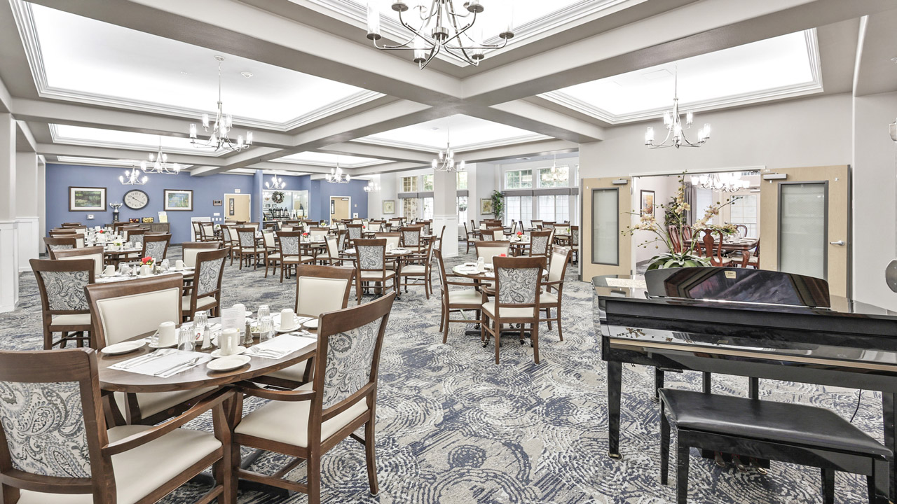 A spacious, elegant dining room with white and wood chairs around neatly set tables, patterned carpet, chandeliers, and a grand piano in the foreground, under a coffered ceiling with bright natural and artificial lighting.