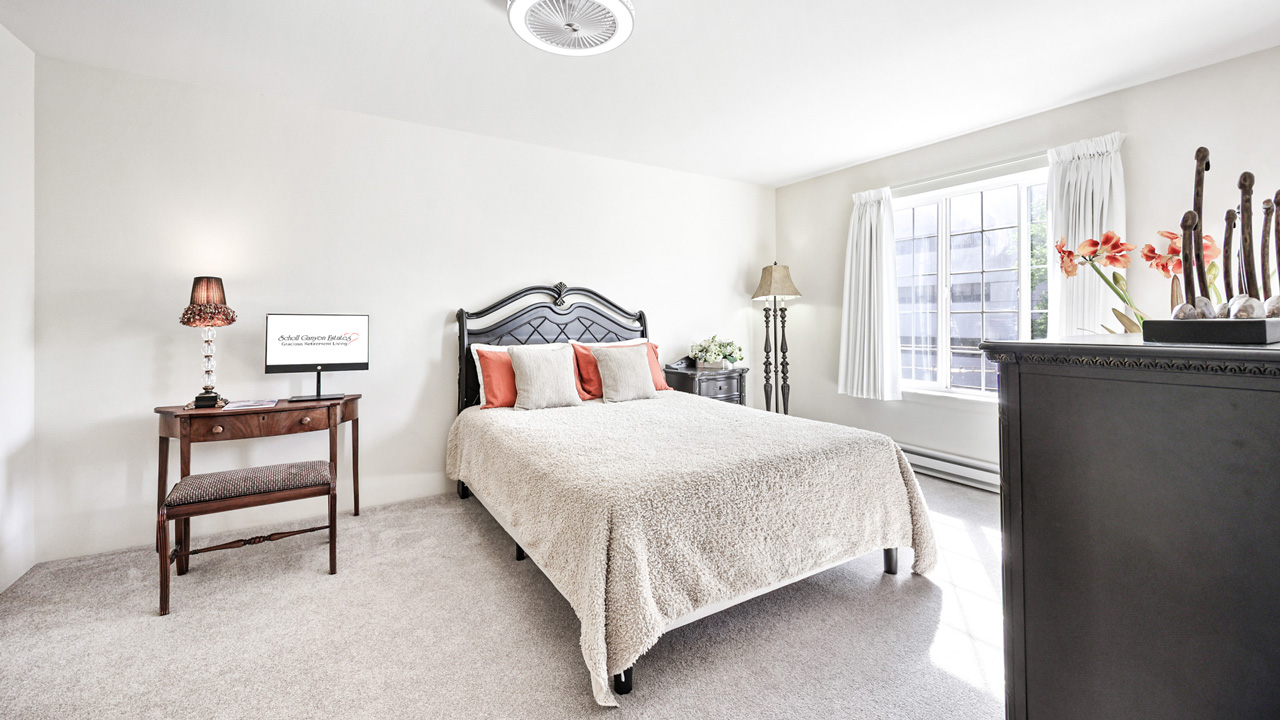 Bright bedroom with a large bed, two orange pillows, a small TV on a wooden table, a bench, and dresser with decor. Sunlight streams through a large window with white curtains. Walls and carpet are light-colored.