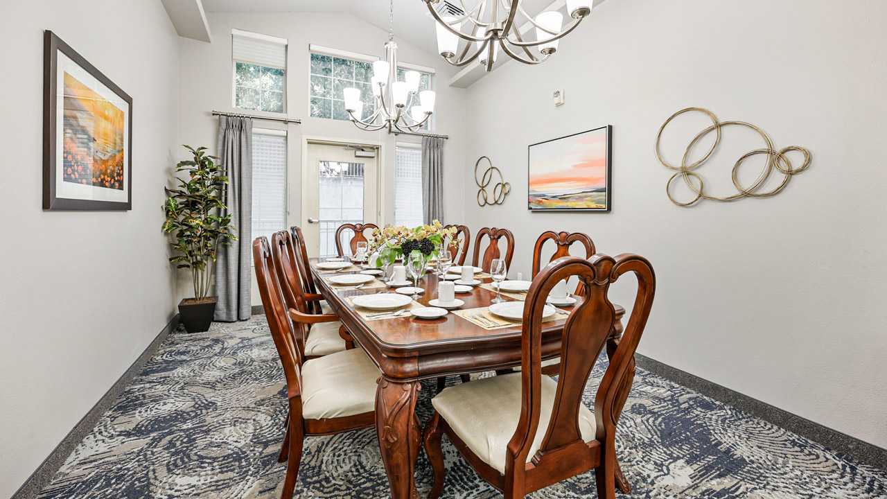A formal dining room with a long wooden table set for eight, cushioned chairs, modern chandeliers, wall art, a potted plant, and large windows with sheer curtains letting in natural light.