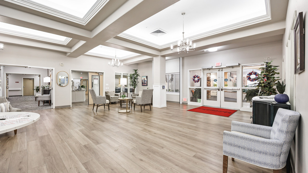 A spacious, well-lit lobby with light wood floors, neutral walls, comfortable seating arrangements, potted plants, and double glass doors decorated with wreaths near an exit. Chandeliers hang from a coffered ceiling.