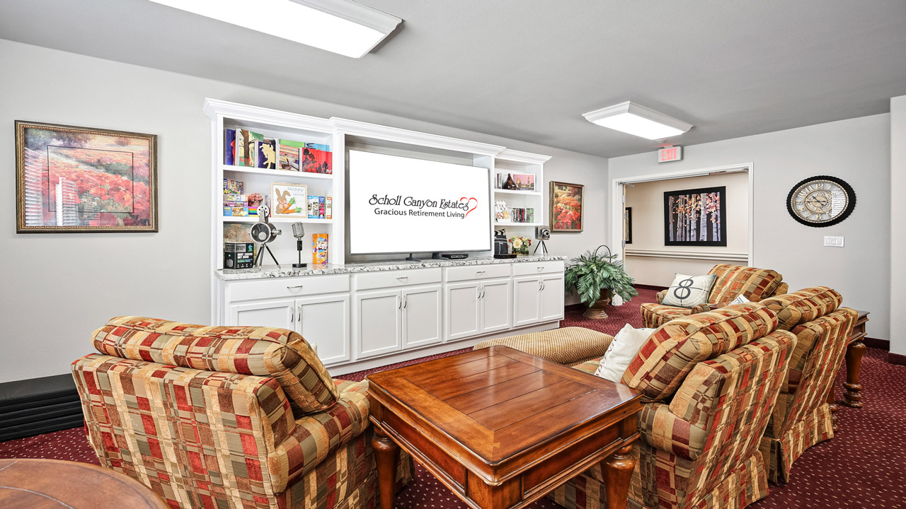 A cozy lounge with patterned armchairs, a large wooden coffee table, built-in white shelves filled with books and decor, a TV displaying “Gracious Retirement Living,” and framed artwork on the cream-colored walls.