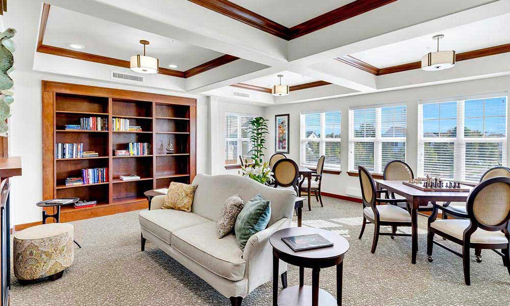 A bright, cozy room featuring a beige sofa with cushions in the center, surrounded by tables and chairs. A large wooden bookshelf filled with books is against the back wall. Windows along the right wall let in ample natural light. A chess set is on one of the tables.