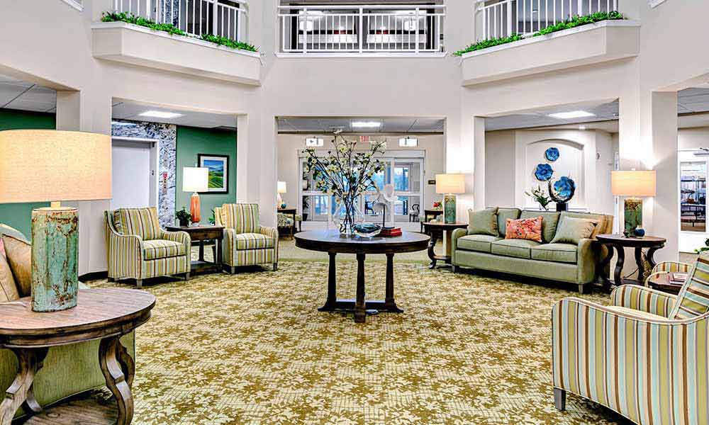 A spacious, well-lit lobby features comfortable seating with striped armchairs and a green sofa. A round wooden table with a flower arrangement sits on a patterned carpet. Lamps and decorative plants add to the welcoming atmosphere, with hallways leading to other areas.