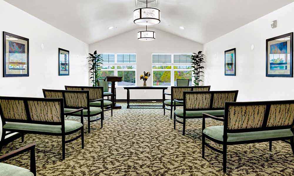 A small, brightly lit room set up for a ceremony with rows of cushioned chairs, a wooden podium, and a table adorned with a flower vase at the front. Potted plants and framed artwork decorate the white walls, and rectangular windows let in natural light.