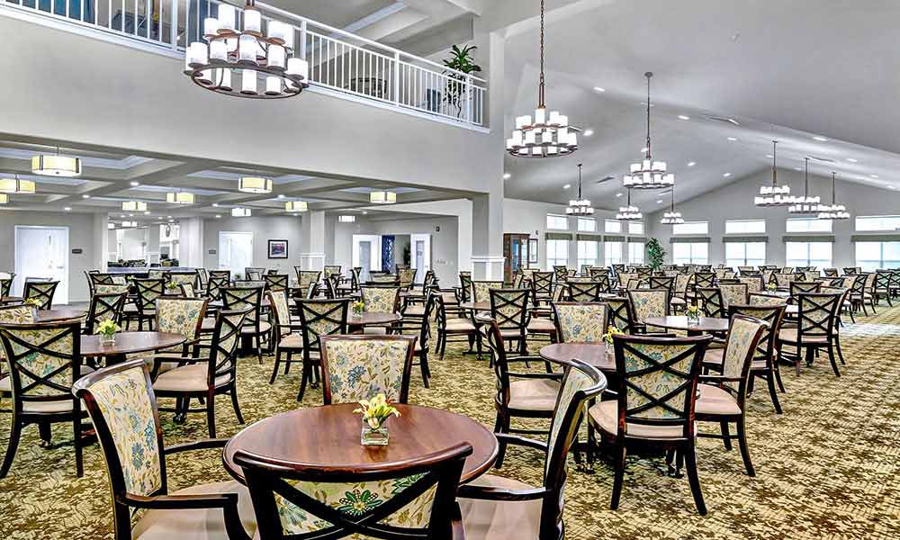 A spacious, well-lit dining area with numerous wooden tables and chairs arranged neatly. The chairs have patterned upholstery. Several chandeliers hang from the high ceiling, and a mezzanine level with a white railing overlooks the space.