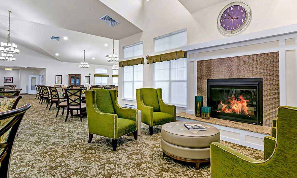 A cozy, well-lit lounge area featuring green upholstered chairs around a round ottoman. A fireplace with a crackling fire adds warmth. The room has carpeted floors, chandeliers, and large windows with blinds. A clock is above the fireplace on the wall.