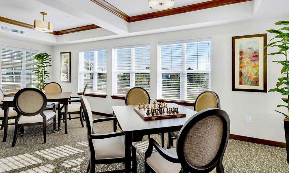 A well-lit room with large windows features a seating area with four chairs around a square table. There is a chessboard on the table. The room also has another table with chairs, potted plants, and colorful paintings on the walls.