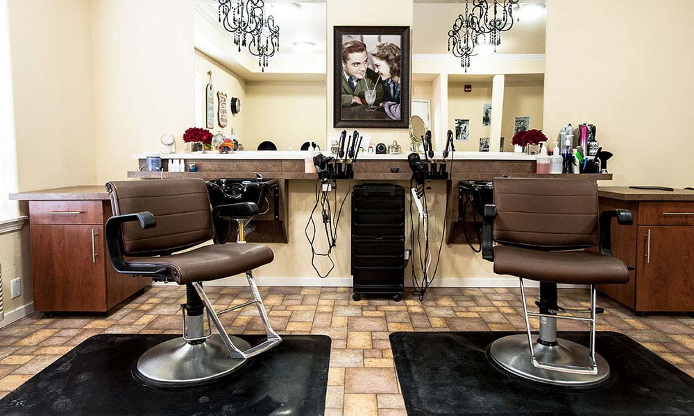 A barbershop scene with two brown barber chairs facing a mirrored wall. Countertops beneath the mirror hold various hair styling tools. A picture of a man and woman from a past era is in the center. The floor is tiled, and two chandeliers hang from the ceiling.