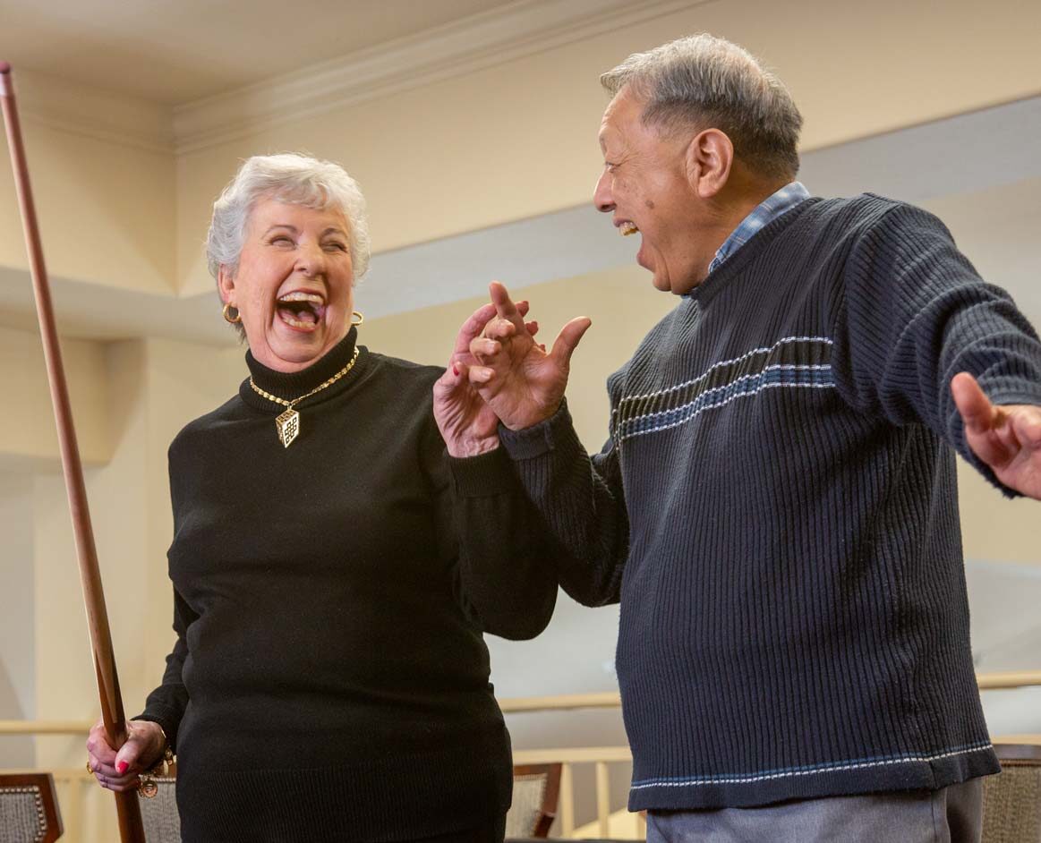An elderly woman and man joyfully laughing together indoors. The woman holds a pool cue, and both are wearing casual sweaters. They appear to be mid-conversation and sharing a light-hearted moment.