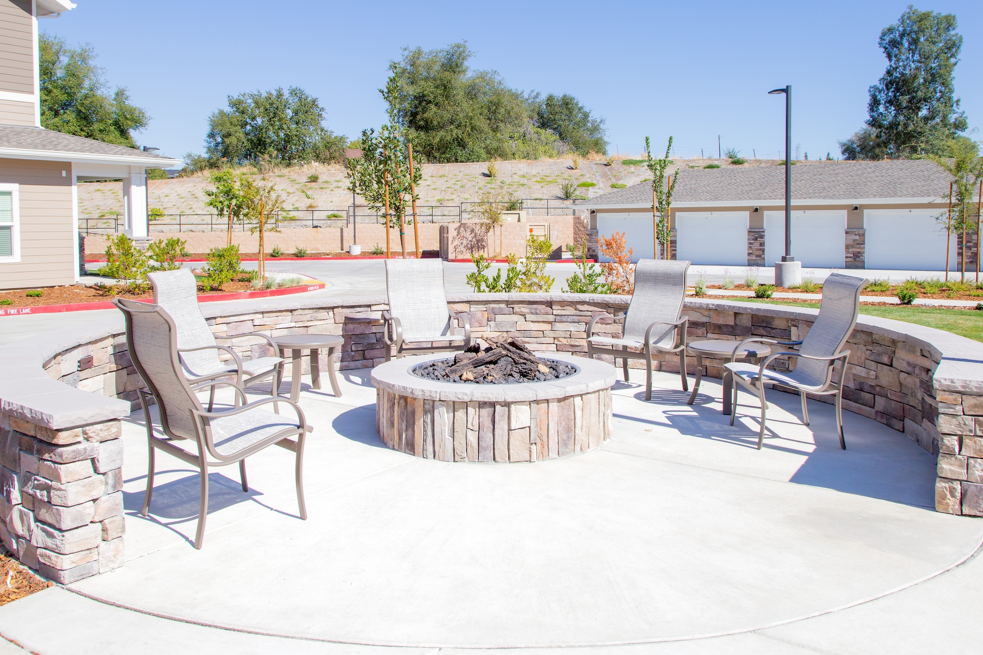 A circular outdoor fire pit area with five metal and mesh chairs is surrounded by a stone wall. The area is set on a concrete patio, with a background of landscaped greenery and a building. Clear blue sky overhead.